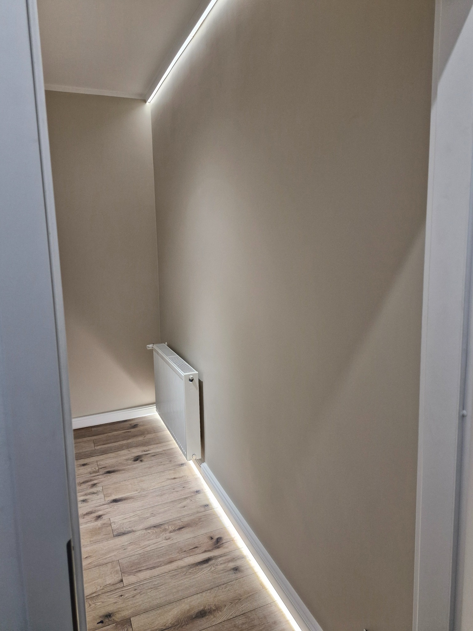 Wnętrze małej garderoby z drewnopodobną podłogą, beżowymi ścianami, grzejnikiem i oświetleniem LED na suficie i przy podłodze.