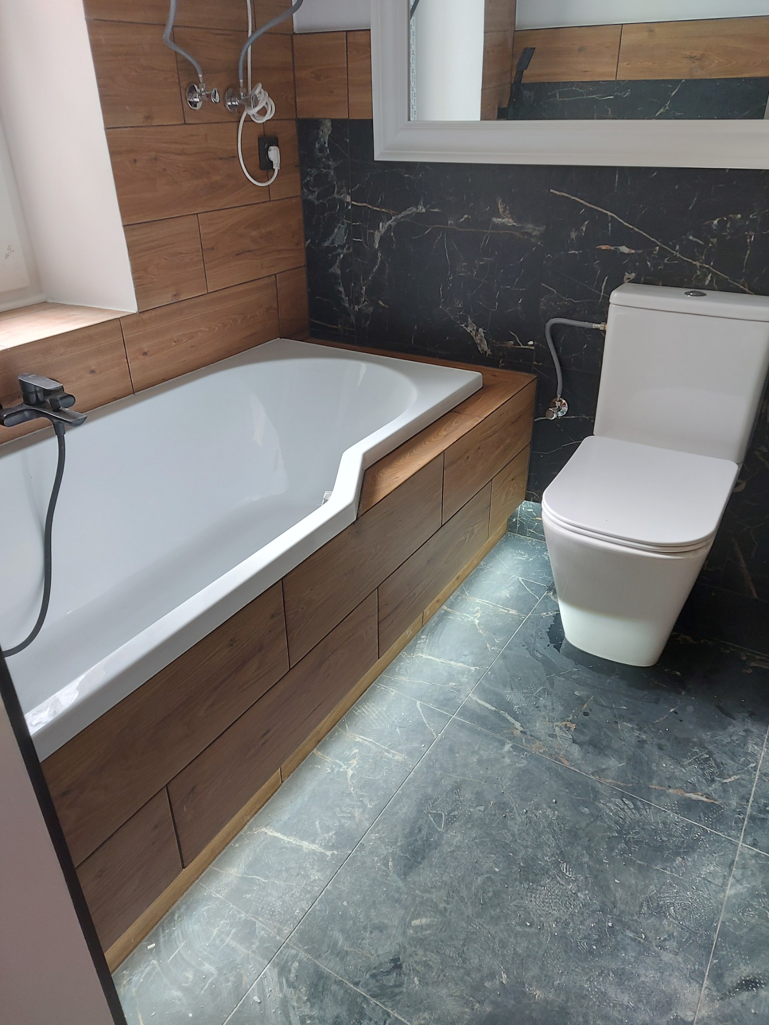 Nowoczesna łazienka z wanną obudowaną drewnopodobnymi panelami, toaletą oraz czarnymi marmurkowymi płytkami na ścianach i podłodze.