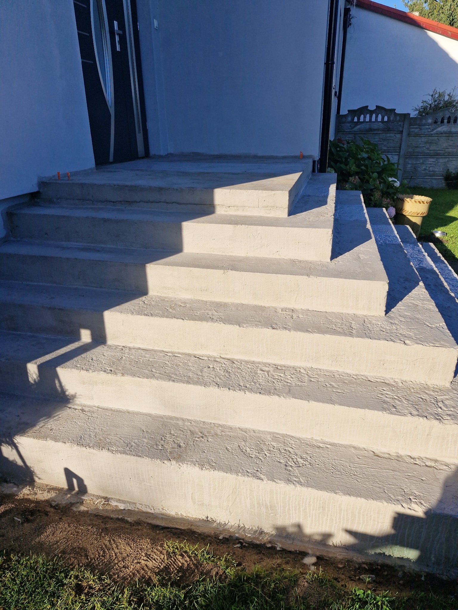 Nowe, betonowe schody wejściowe do domu, prowadzące do drzwi wejściowych. Widoczne ślady świeżego betonu i otoczenie z zielenią.