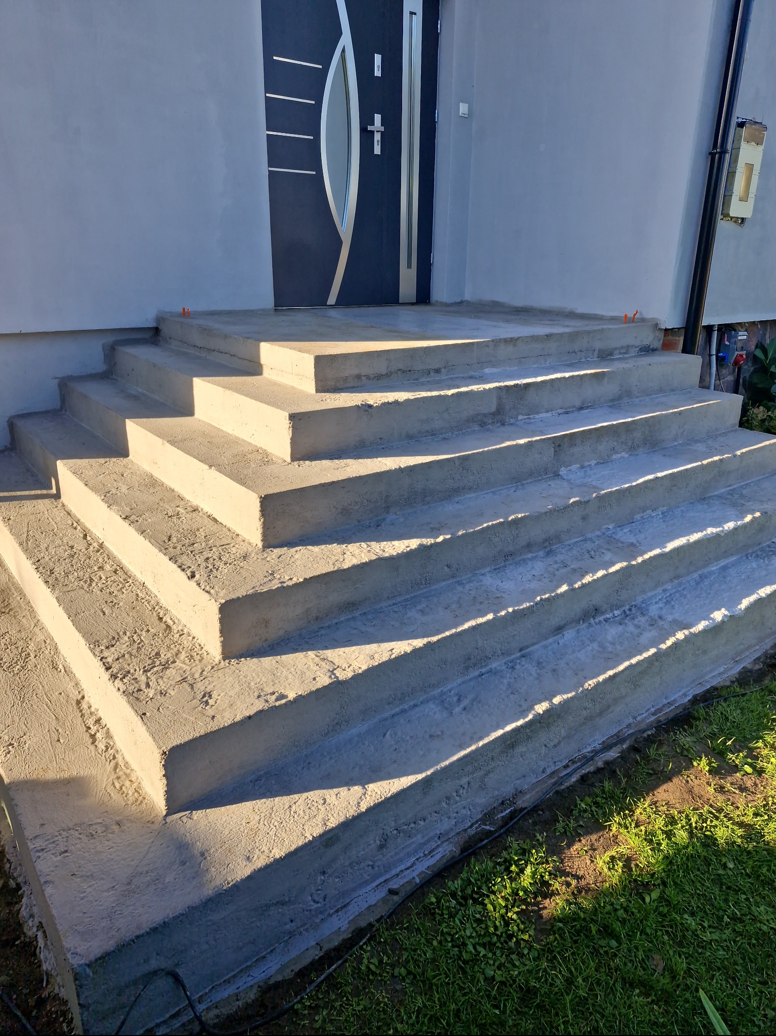 Nowe, betonowe schody wejściowe prowadzące do domu z nowoczesnymi drzwiami. Widoczne świeże wykończenie i detale architektoniczne. Słoneczny dzień.