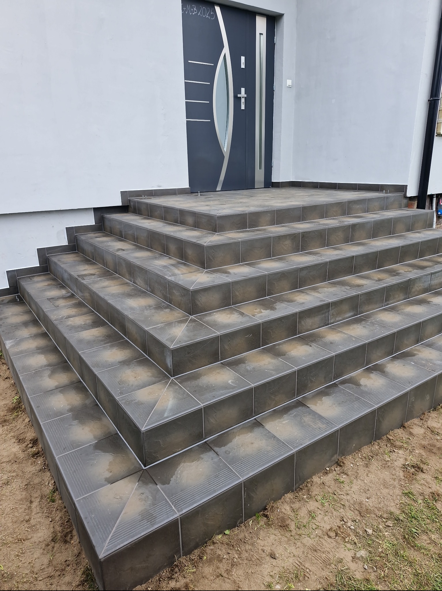 Wykończone płytkami schody wejściowe do domu, z szarą stolarką drzwiową i fragmentem elewacji. Widoczne dokładne ułożenie płytek na stopniach i podstopnicach.