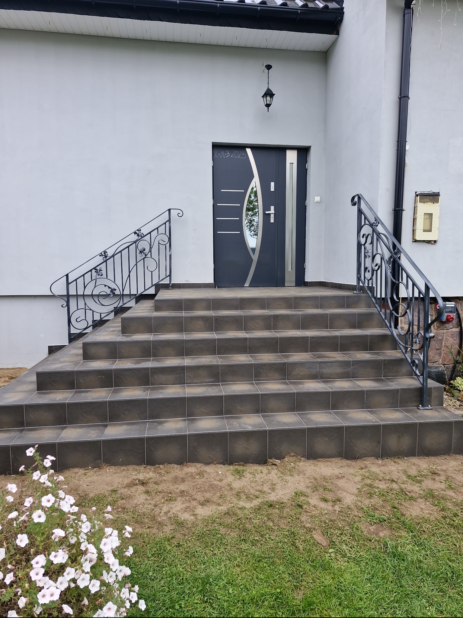 Zewnętrzne schody wejściowe wyłożone brązowymi płytkami, z ozdobną, metalową balustradą. Szare ściany budynku i nowoczesne drzwi wejściowe w tle. Kwiaty w dolnej części kadru.