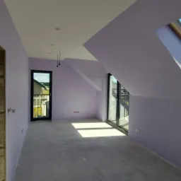 Poddasze po malowaniu na kolor liliowy, widoczne okno dachowe z widokiem na błękitne niebo, surowa ościeżnica drzwiowa i gładka wylewka na podłodze.