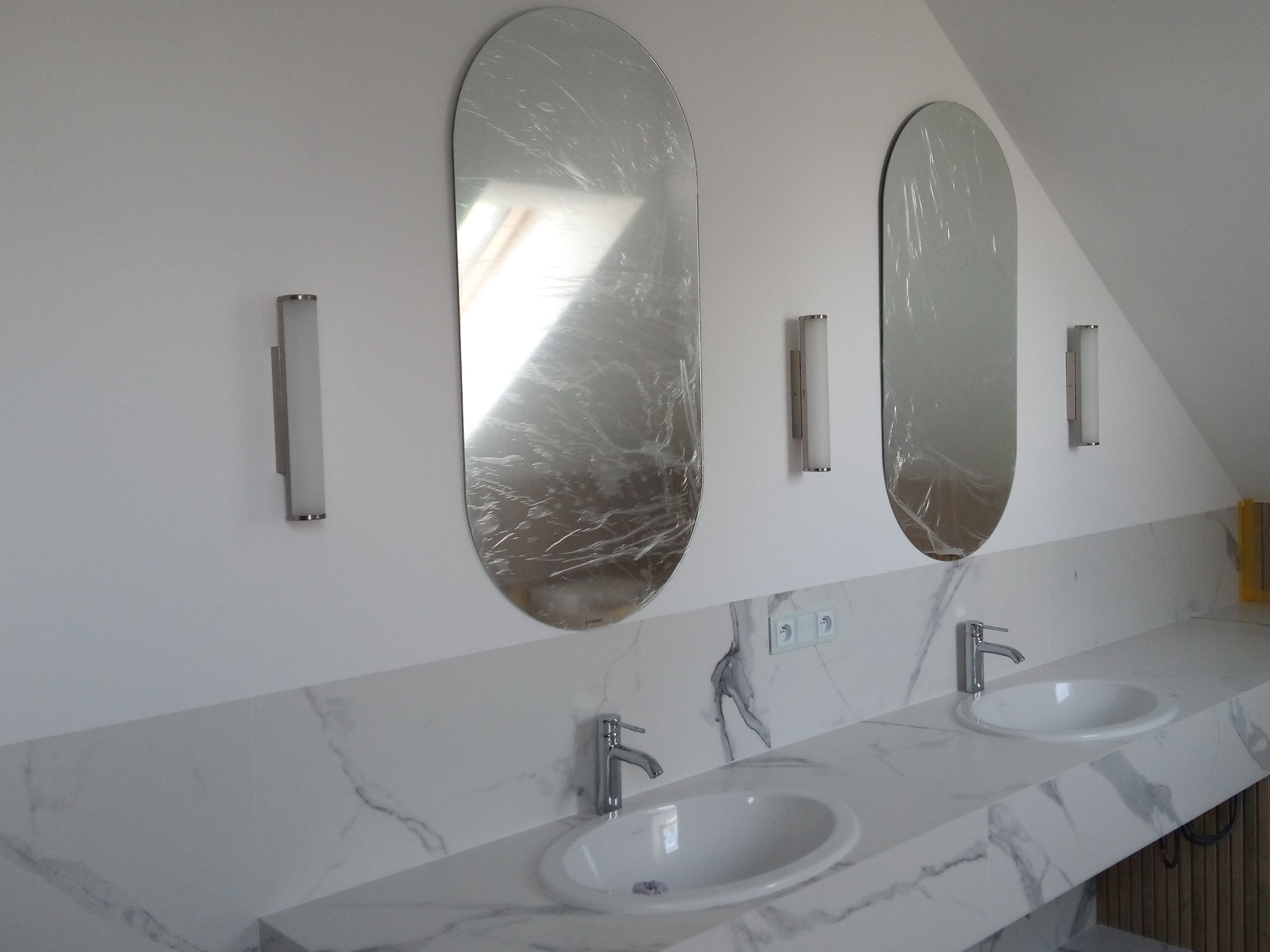 Nowoczesna łazienka na poddaszu z dwoma owalnymi umywalkami i lustrami w folii ochronnej, oświetlona podłużnymi lampami ściennymi.