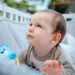 Niemowlę z niebieskimi oczami leżące w łóżeczku, trzymające zabawkę misia i spoglądające w górę, z zielenią widoczną w tle.