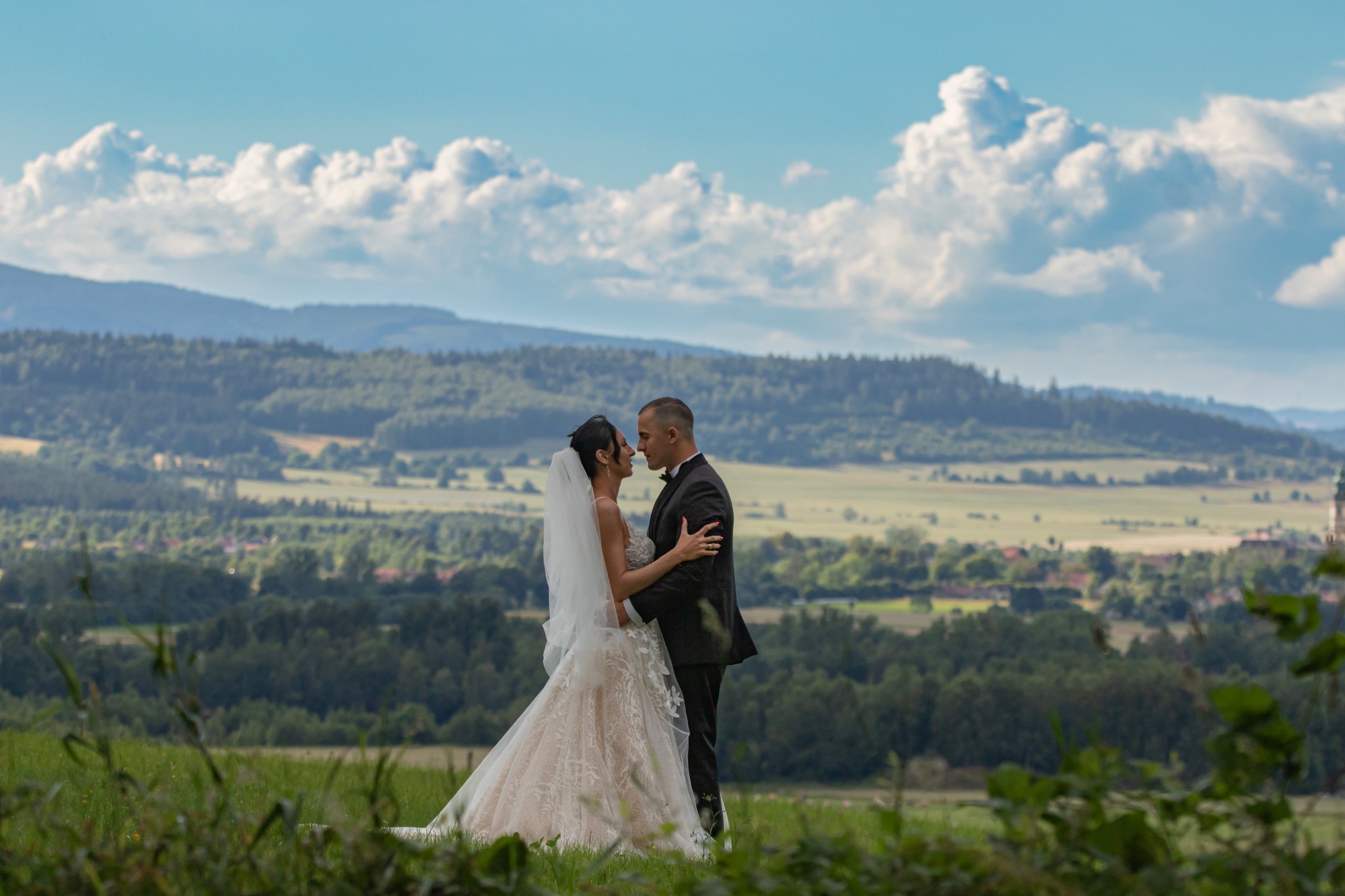 Para młoda w objęciach na tle malowniczego krajobrazu z górami i chmurami, panna młoda w sukni z koronkowymi aplikacjami, pan młody w czarnym smokingu.