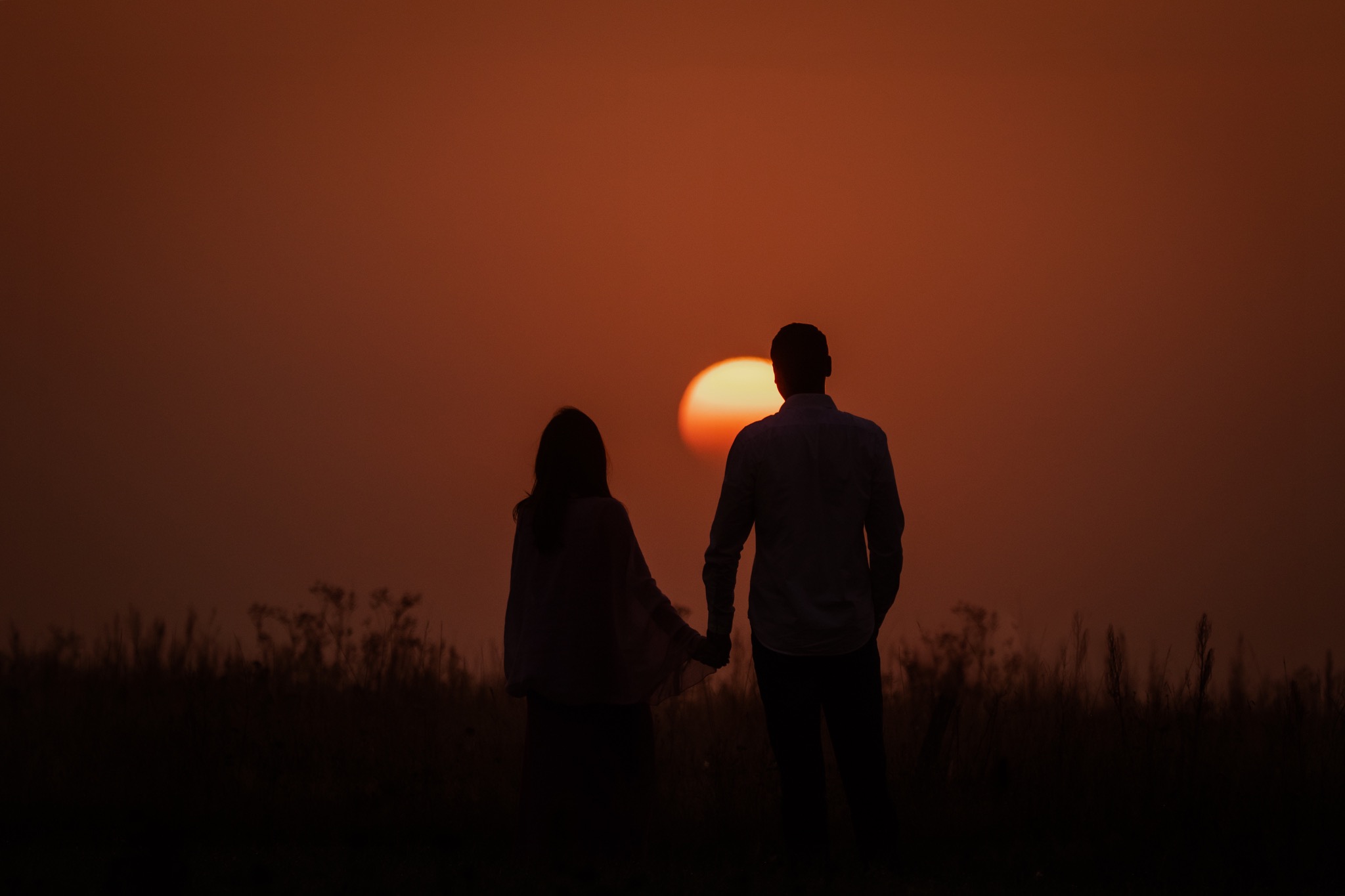Para trzymająca się za ręce, sylwetki na tle zachodzącego słońca, widok z tyłu, wysoka trawa na pierwszym planie.