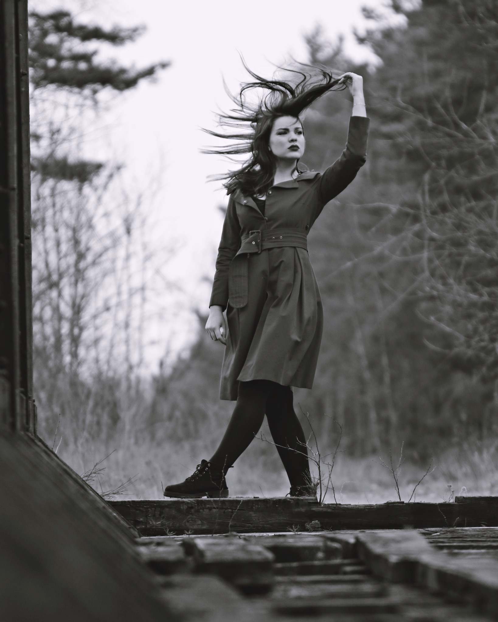 Czarno-białe zdjęcie kobiety w płaszczu i botkach, stojącej na drewnianej konstrukcji z podniesioną ręką we włosach, z zamazanym tłem drzew.