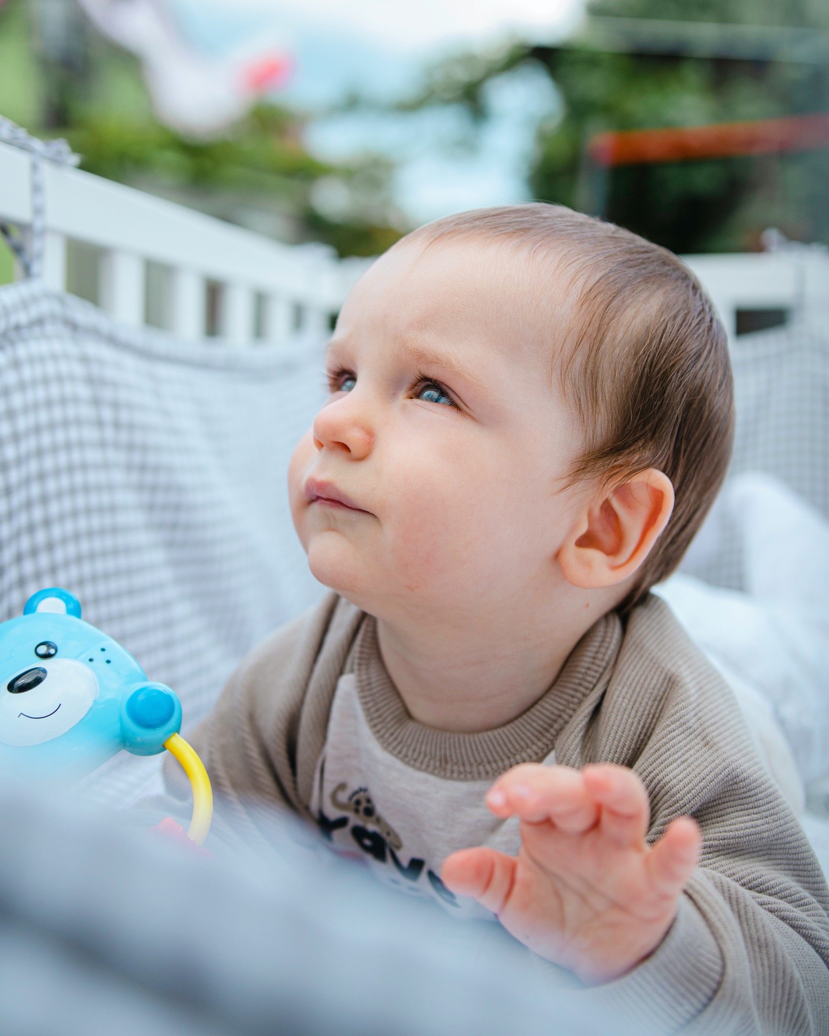 Niemowlę z niebieskimi oczami leżące w łóżeczku, trzymające zabawkę misia i spoglądające w górę, z zielenią widoczną w tle.