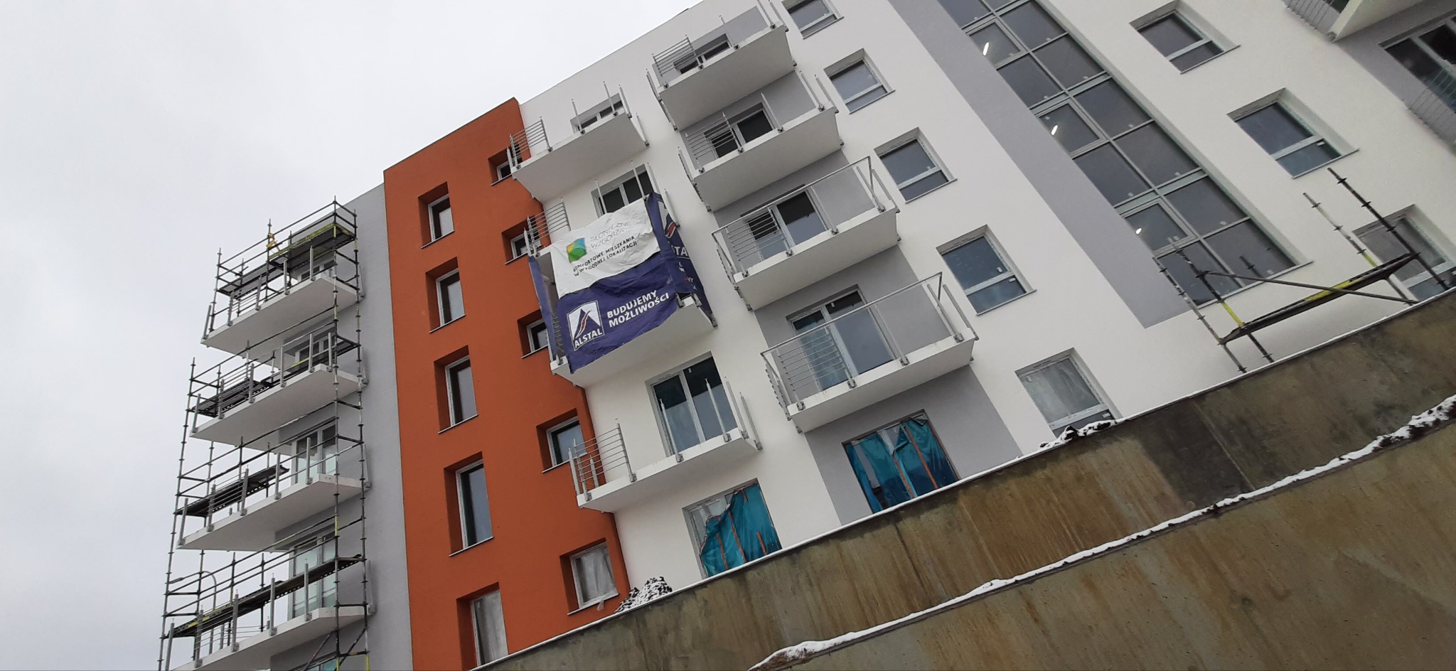 Nowo wybudowany budynek wielorodzinny w Gdańsku z pomarańczową ścianą i balkonami, na których wisi baner reklamowy firmy deweloperskiej; widoczne rusztowania.