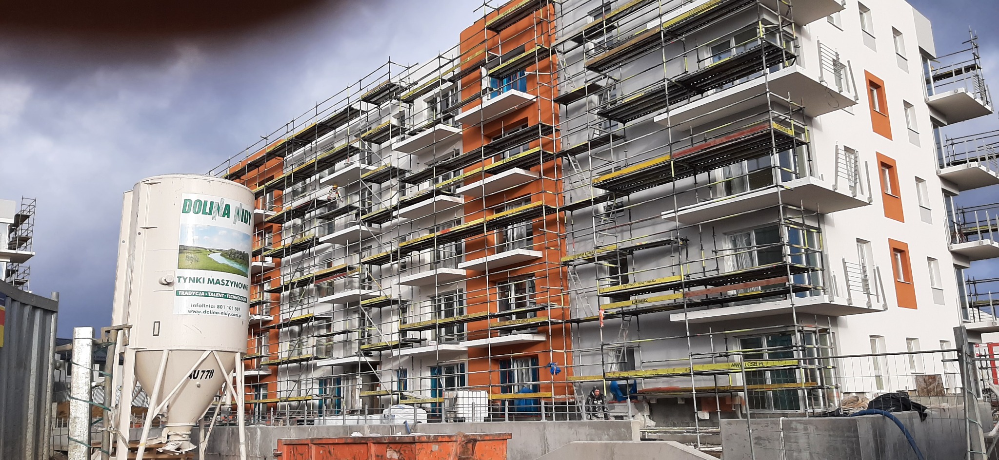 Wielopiętrowy budynek w trakcie renowacji elewacji, otoczony rusztowaniami, z widocznym silosie na tynk maszynowy firmy Dolina Nidy.