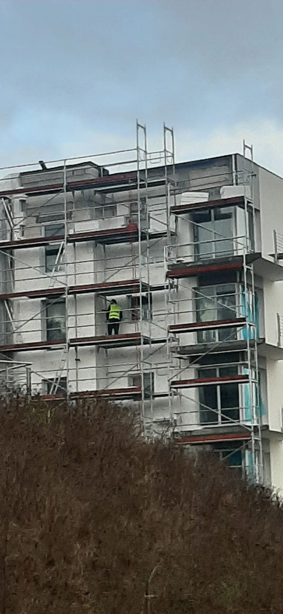 Pracownik w żółtej kamizelce na rusztowaniu przy białym budynku wielorodzinnym, prawdopodobnie podczas prac elewacyjnych.