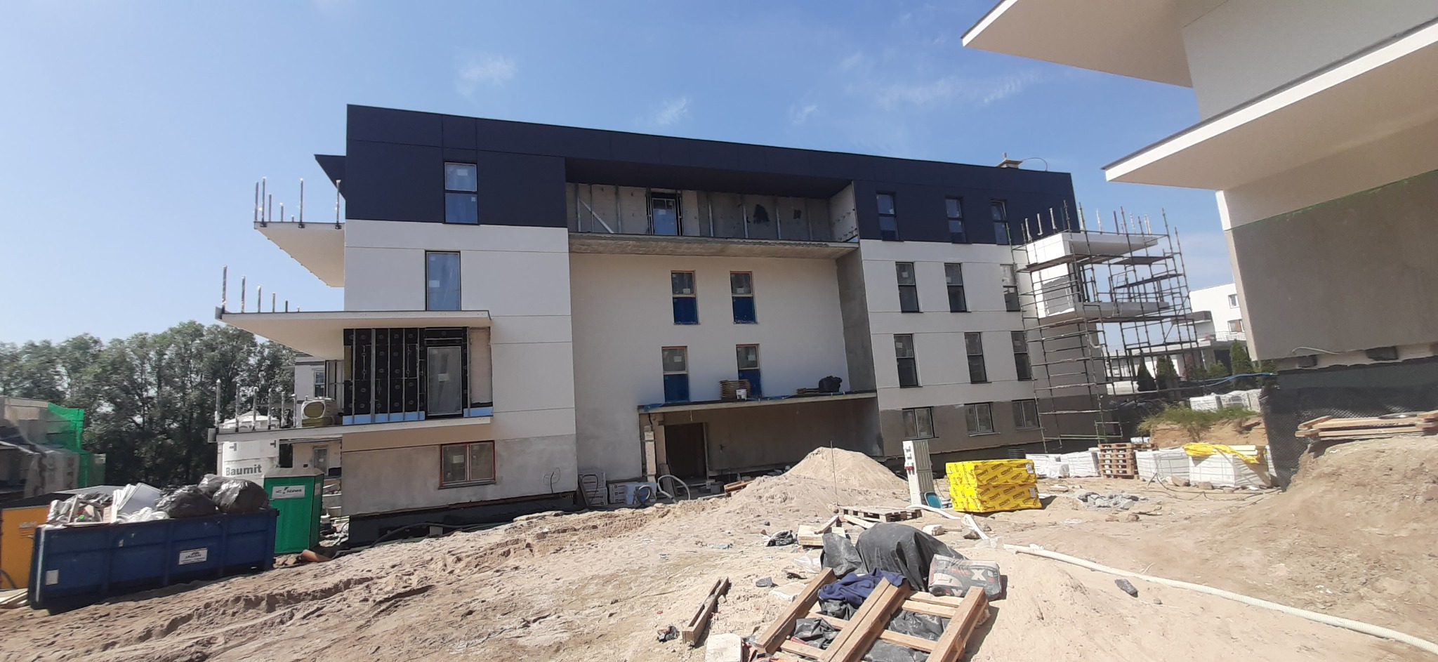 Nowoczesny budynek wielorodzinny w trakcie budowy, widoczne rusztowania, niezabezpieczone otwory okienne i materiały budowlane na terenie budowy.