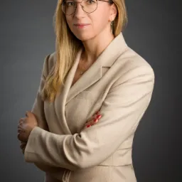 Portret kobiety w beżowym garniturze, okularach i z blond włosami, na szarym tle, z założonymi rękami.