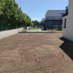 Przygotowany teren pod trawnik, wyrównana ziemia przed nowymi domami, widoczna siatka ogrodzeniowa i droga z drzewami w tle.