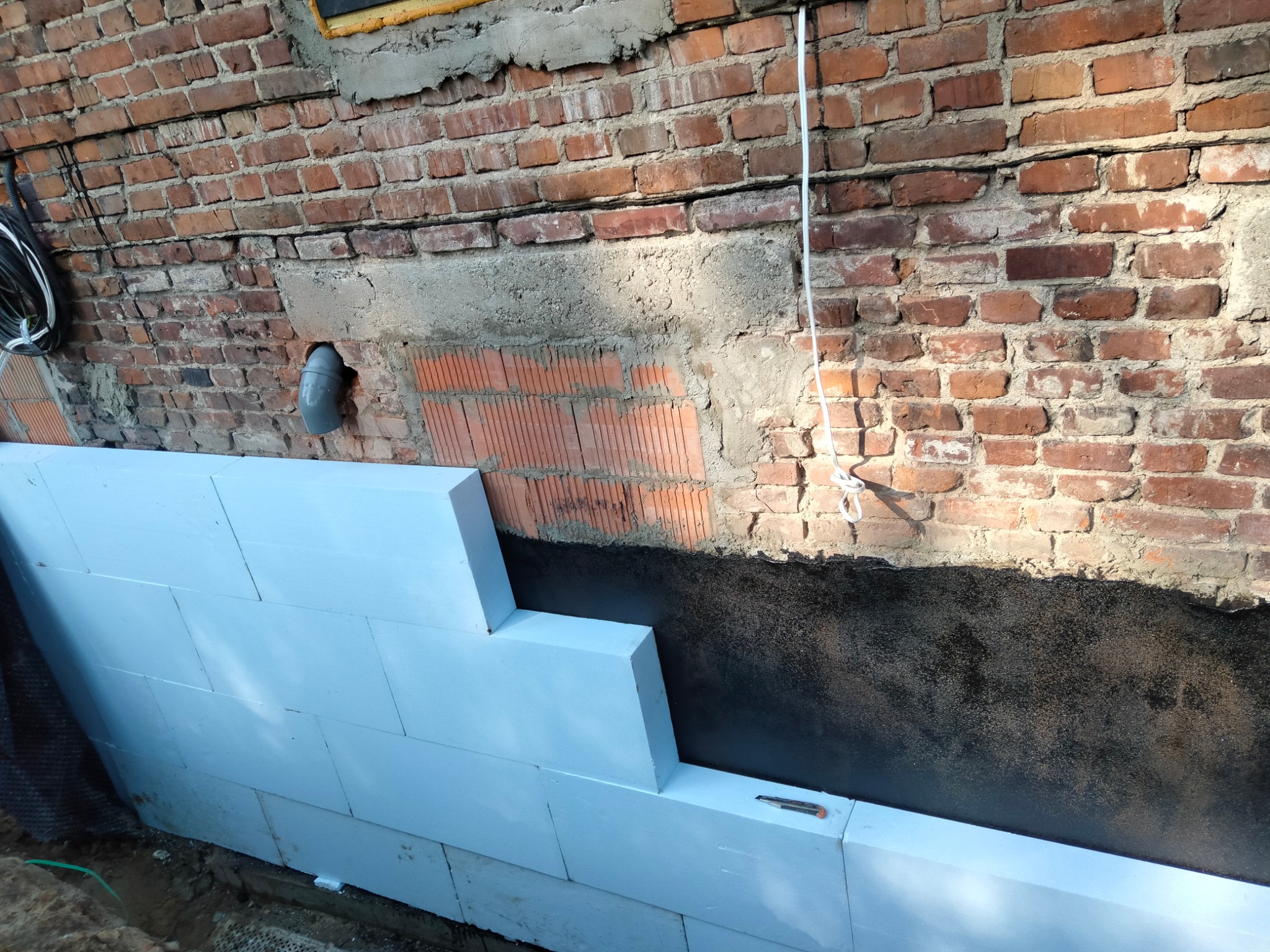 Fundament budynku z cegły zabezpieczony czarną izolacją przeciwwilgociową i ocieplony styropianem, na którym leży nóż do cięcia.