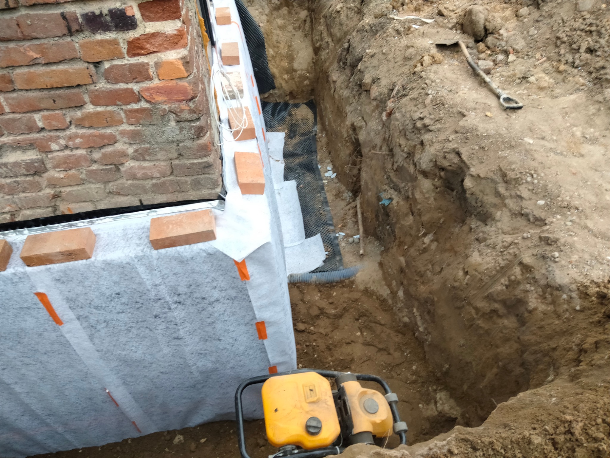 Wykop pod fundament z izolacją pionową zabezpieczoną geowłókniną, ubijarką wibracyjną i łopatą w tle.
