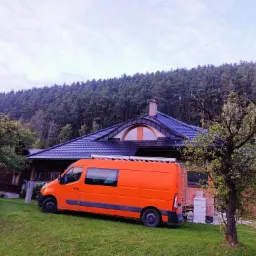 Pomarańczowy samochód dostawczy z drabiną na dachu zaparkowany na trawie przed domem z ciemnym dachem i widocznym kominem na tle lasu.