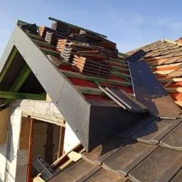 Wymiana dachu z dachówki ceramicznej na budynku w trakcie budowy, widoczne elementy więźby dachowej, obróbki blacharskie i stos dachówek na dachu.