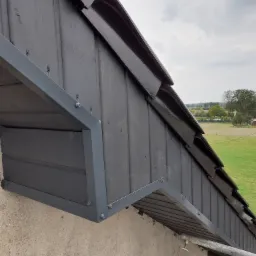 Szczegółowe ujęcie pomalowanej na czarno drewnianej elewacji szczytowej dachu z widocznymi obróbkami blacharskimi w kolorze szarym, na tle zielonego krajobrazu.