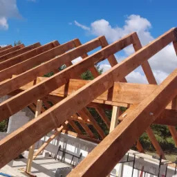 Konstrukcja drewnianej więźby dachowej z widocznym niebem i chmurami, elementy rusztowania i pustaków na budowie.