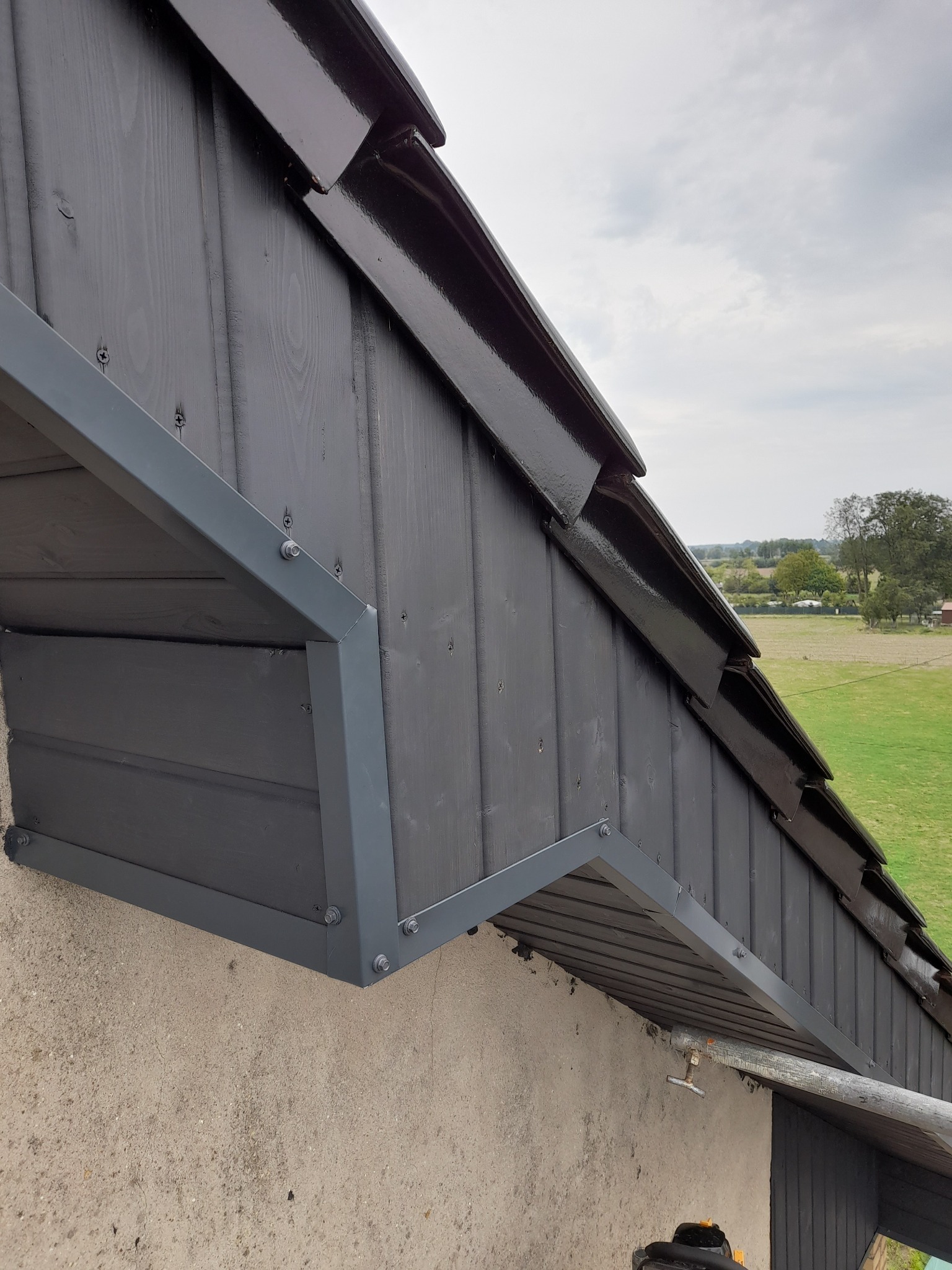 Szczegółowe ujęcie pomalowanej na czarno drewnianej elewacji szczytowej dachu z widocznymi obróbkami blacharskimi w kolorze szarym, na tle zielonego krajobrazu.