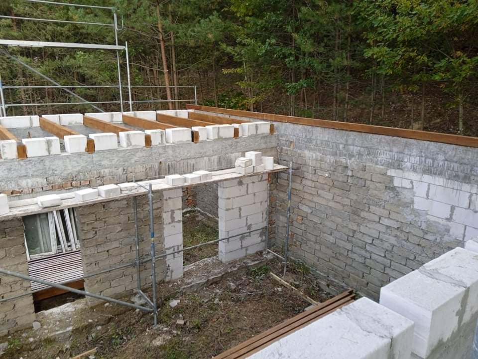 Budowa domu w trakcie: ściany z białego betonu komórkowego, drewniane belki stropowe, rusztowanie w tle, widok z góry na fundamenty.