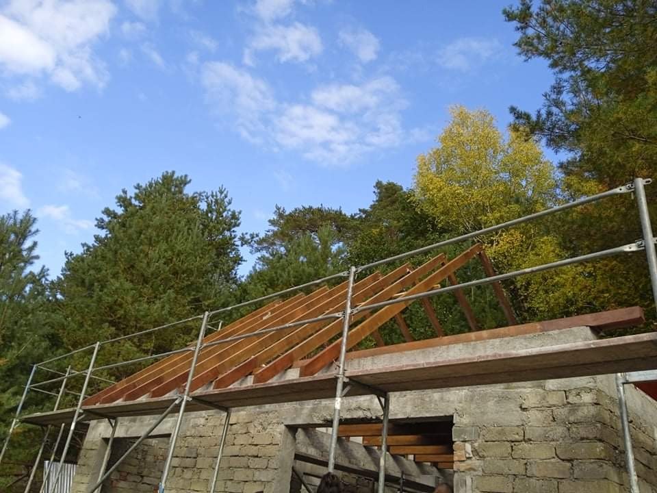 Budowa dachu z drewnianą konstrukcją na murowanym budynku, widoczne rusztowania i drzewa w tle pod błękitnym niebem.