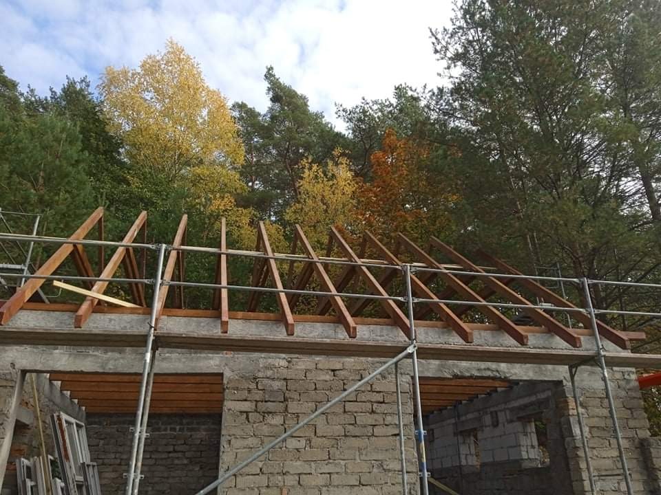 Wznoszenie drewnianej konstrukcji dachu na murowanym budynku, widoczne rusztowania i jesienne drzewa w tle.