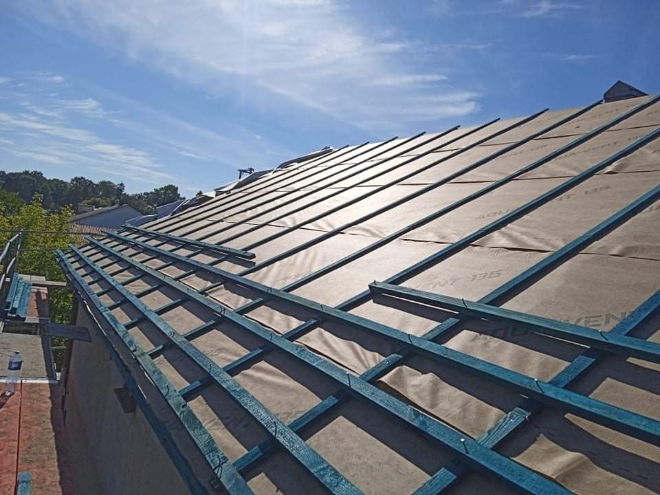 Widok z góry na konstrukcję dachu w trakcie budowy, z widoczną membraną i niebieskimi łatami, na tle błękitnego nieba.