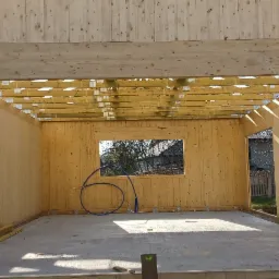 Wnętrze surowego domu z drewnianych paneli, widoczne elementy konstrukcyjne dachu i okno z widokiem na zewnątrz, na betonowej podłodze zwinięty niebieski przewód.