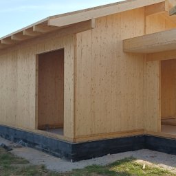 Darek - Surowy stan domu szkieletowego z jasnego drewna, widoczne otwory okienne i drzwiowe, czarna izolacja fundamentów, w tle hałda ziemi.