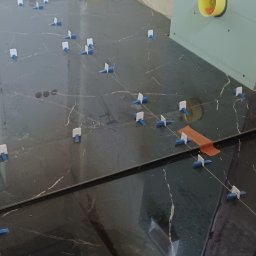 Darek - Układanie ciemnych płytek z marmurowym wzorem, widoczne kliny i krzyżyki dystansowe, fragment ściany z żółtym wlotem wentylacyjnym.