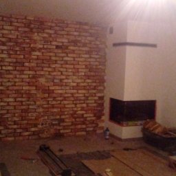 Darek - Ściana z cegły dekoracyjnej w trakcie montażu, widoczny kominek, narzędzia glazurnicze i odkurzacz, etap prac wykończeniowych w pomieszczeniu.