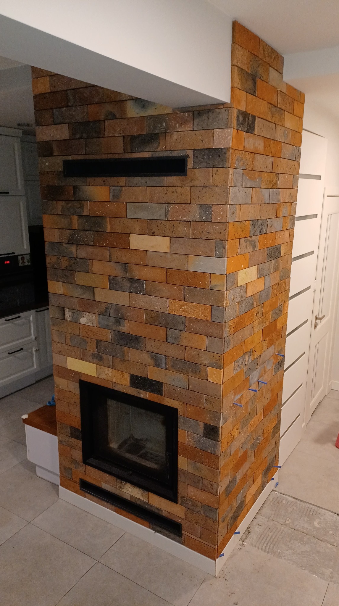 Wykończony kominek obłożony cegłą klinkierową w odcieniach brązu, pomarańczy i szarości, z czarnym wkładem kominkowym i kratkami wentylacyjnymi, widoczne niebieskie kliny dystansowe na dole ściany.