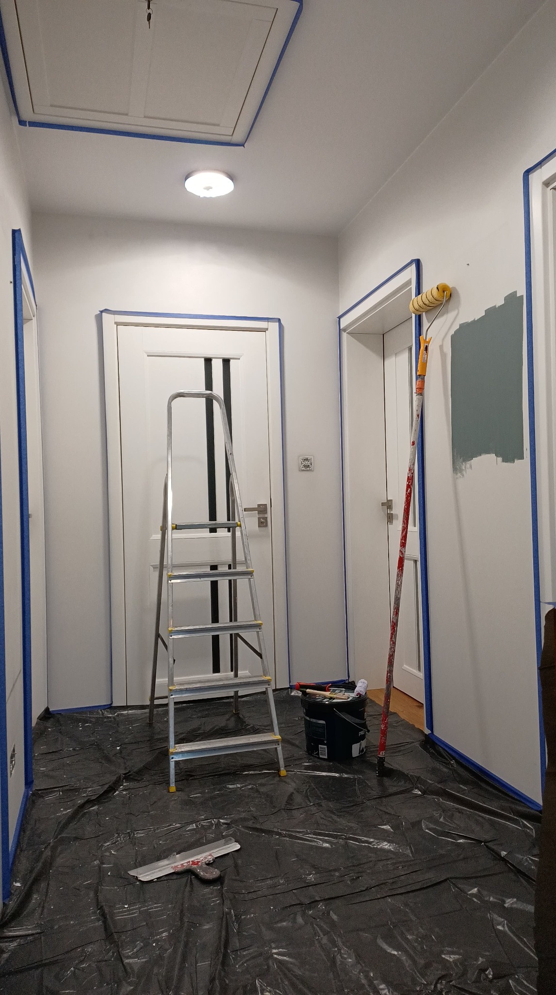 Korytarz w trakcie malowania, zabezpieczony folią i taśmą malarską, widoczna drabina, wałek i częściowo pomalowana ściana na szaro.