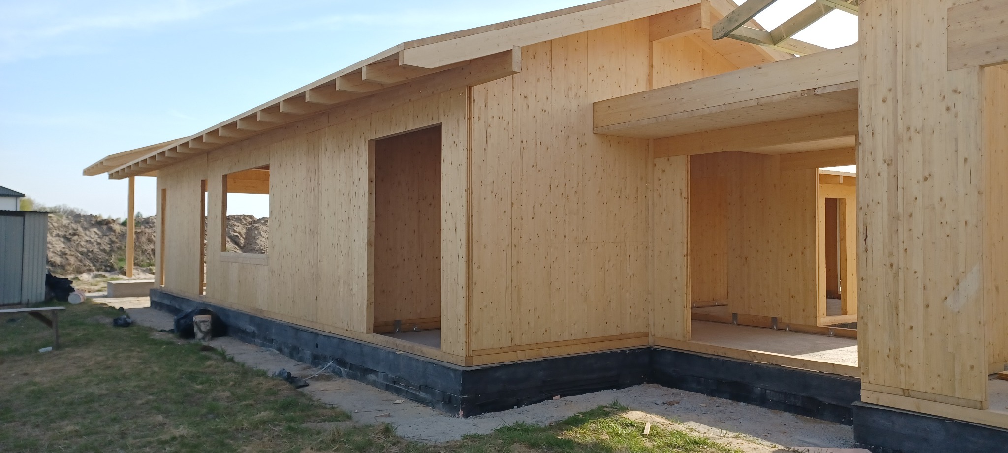 Surowy stan domu szkieletowego z jasnego drewna, widoczne otwory okienne i drzwiowe, czarna izolacja fundamentów, w tle hałda ziemi.