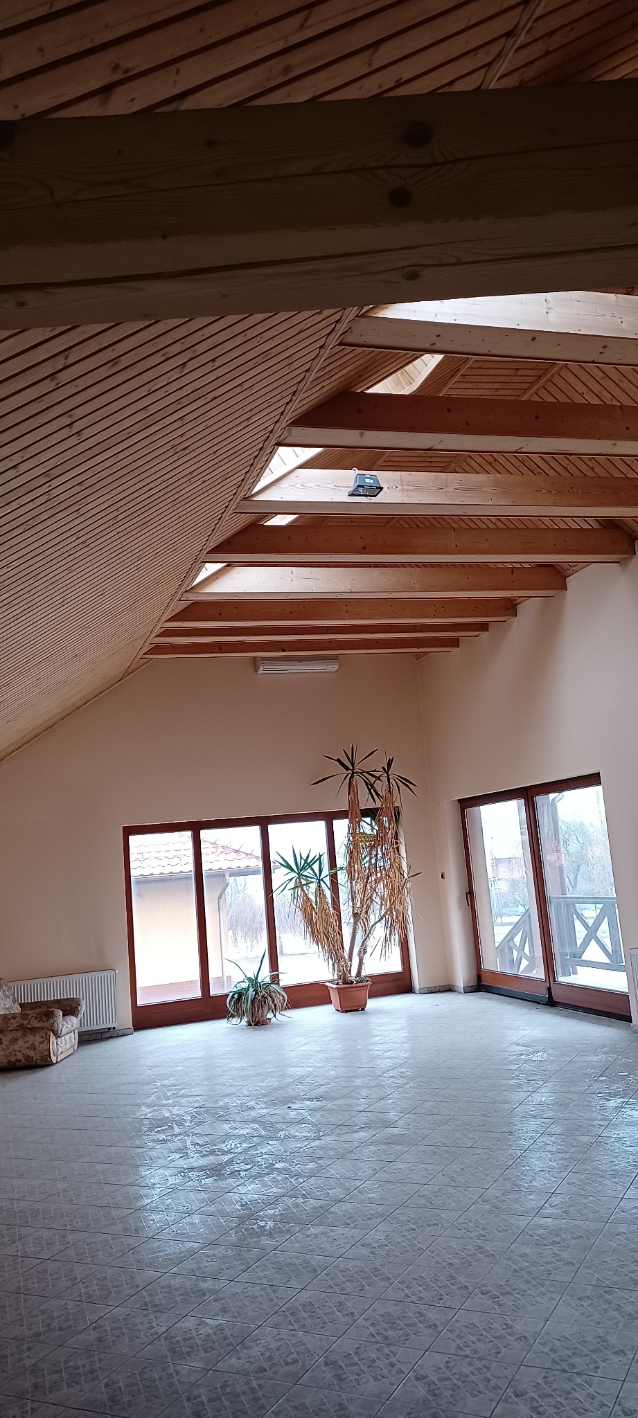 Przestronne poddasze z drewnianym wykończeniem sufitu, świeżo położona szara glazura podłogowa z wzorem, duże okna balkonowe i roślina doniczkowa w słoneczny dzień.