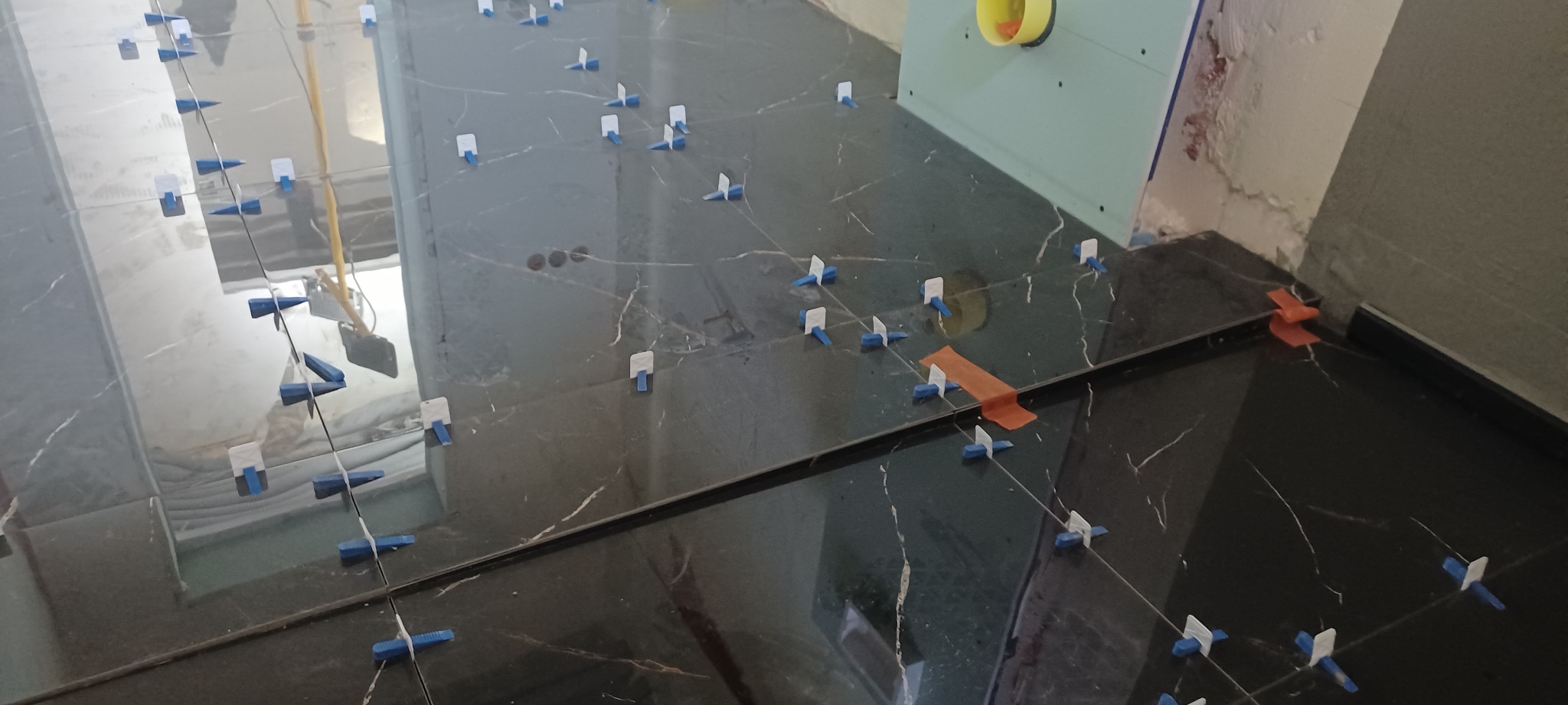 Układanie ciemnych płytek z marmurowym wzorem, widoczne kliny i krzyżyki dystansowe, fragment ściany z żółtym wlotem wentylacyjnym.