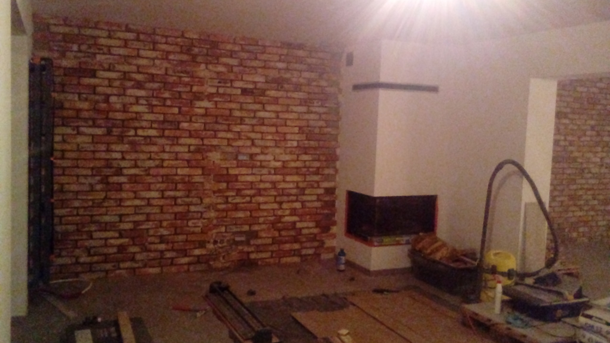 Ściana z cegły dekoracyjnej w trakcie montażu, widoczny kominek, narzędzia glazurnicze i odkurzacz, etap prac wykończeniowych w pomieszczeniu.