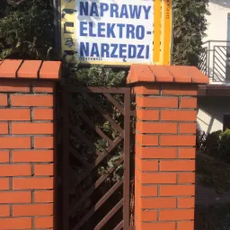 Tablica reklamowa zakładu naprawy elektronarzędzi umieszczona na ceglanym słupku ogrodzenia, widoczne ikony elektronarzędzi i numer telefonu.