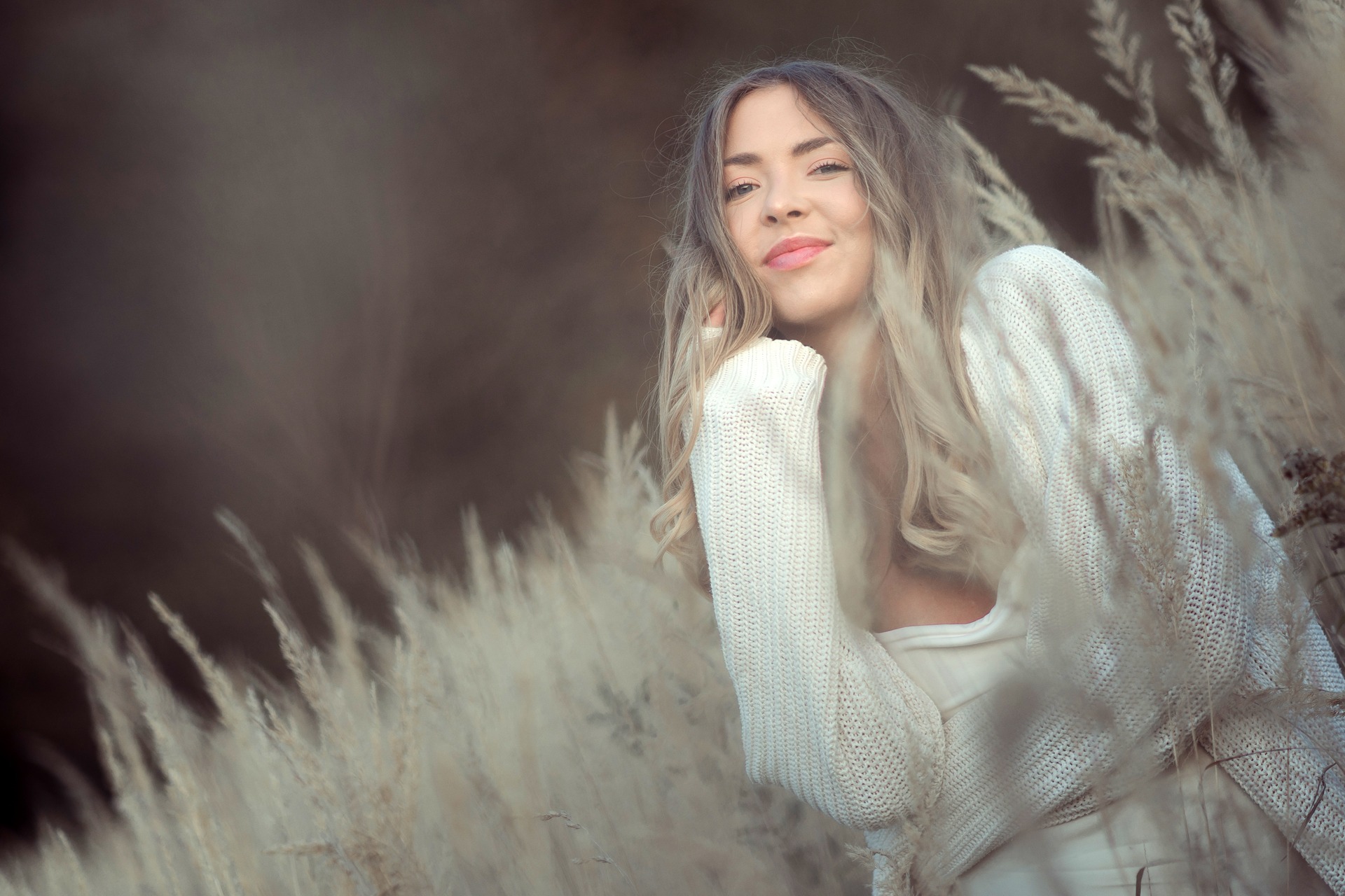 Portret uśmiechniętej kobiety w białym swetrze, pozującej w wysokiej trawie w jesiennej scenerii, z delikatnym oświetleniem podkreślającym rysy twarzy.