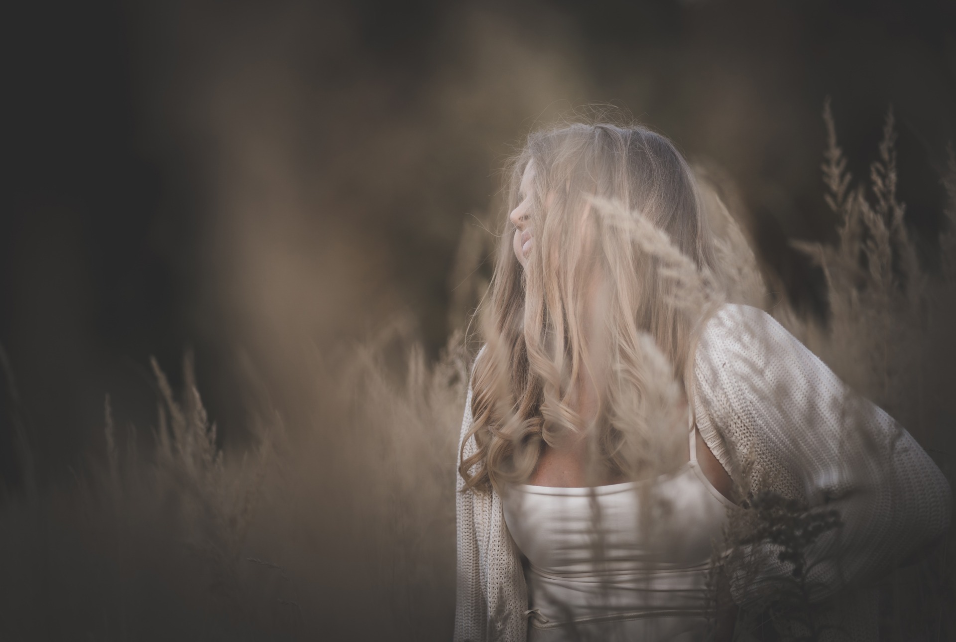 Portret kobiety z profilu, z rozpuszczonymi blond włosami zasłaniającymi twarz, w otoczeniu wysokich traw w jesiennej tonacji.