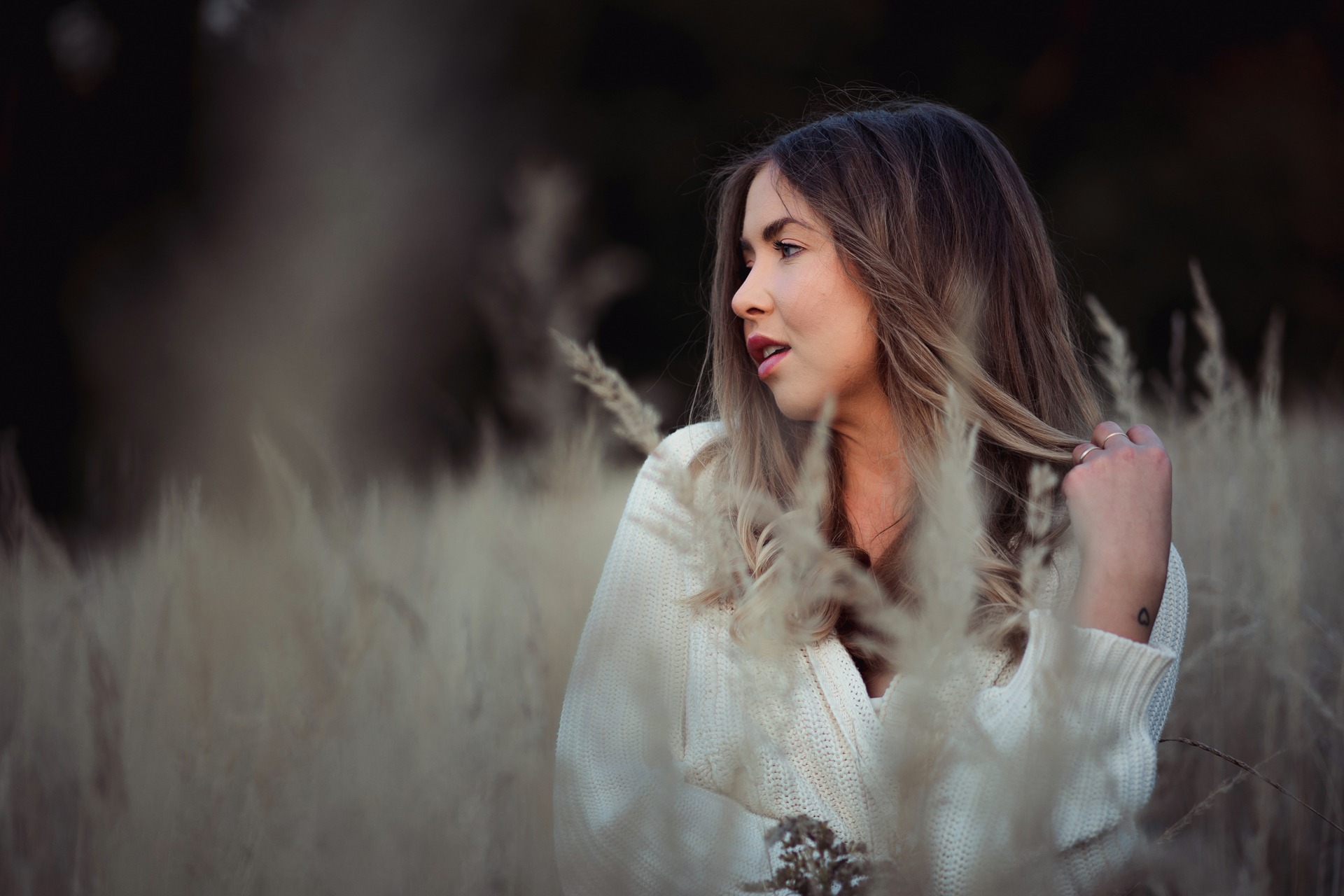 Portret kobiety w białym swetrze, stojącej w wysokiej trawie, z delikatnym rozmyciem na pierwszym planie i ciemnym tłem, spoglądającej w lewo.