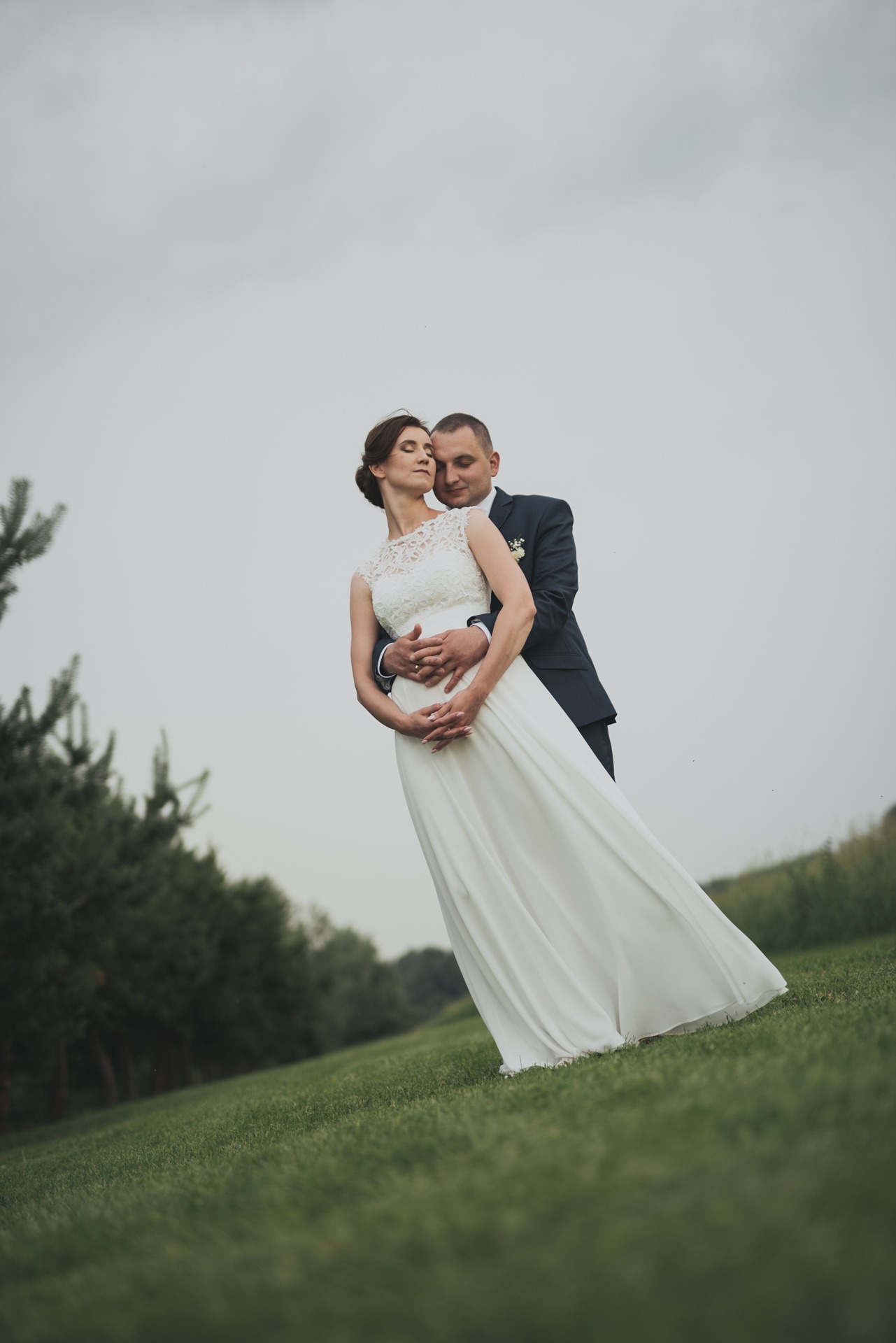 Para młoda na zielonym wzgórzu, panna młoda w koronkowej sukni z zamkniętymi oczami, pan młody obejmuje ją w granatowym garniturze.