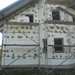 Dom w trakcie prac związanych z izolacją termiczną ścian zewnętrznych. Widoczne płyty styropianowe przyklejone do elewacji, częściowo zamaskowane zaprawą, oraz rusztowanie. Na rusztowaniu leży...