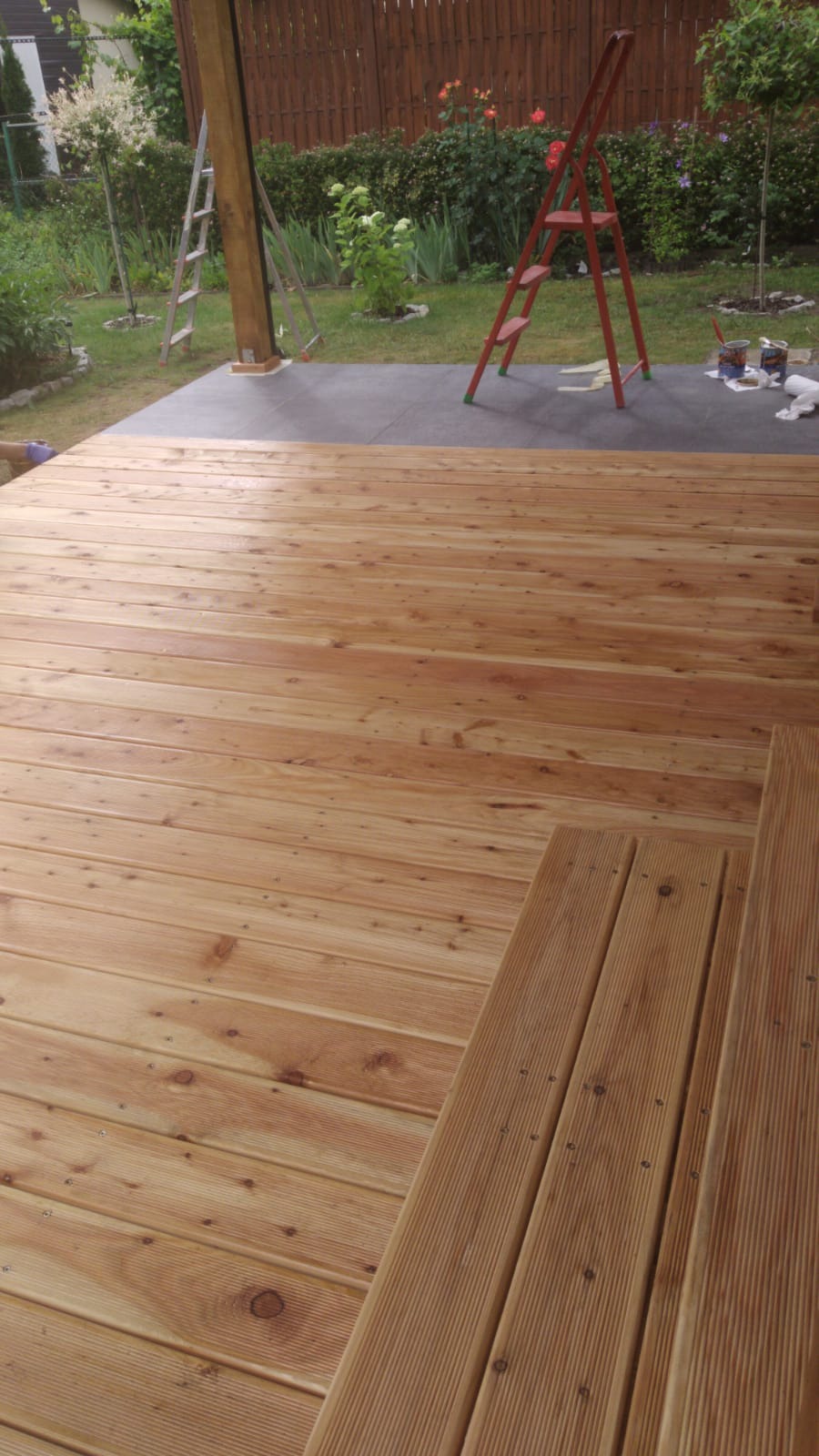 Wykończony drewniany taras z widocznymi schodami, w tle trawnik, drabina i narzędzia malarskie.