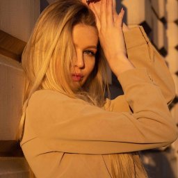 Katerina Bila - Portret kobiety z blond włosami, w beżowej marynarce i czarnym pasku, opierającej się o ścianę w ciepłym, zachodzącym słońcu, z dłońmi uniesionymi nad głową.