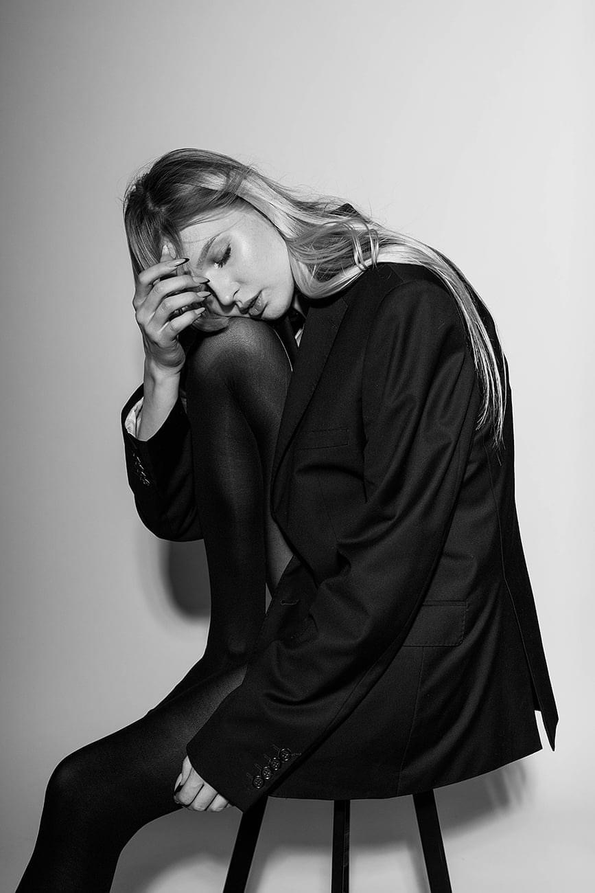 Czarno-białe zdjęcie kobiety w oversize'owej marynarce siedzącej na stołku, z głową opartą na dłoni, kolana podciągnięte pod brodę, w ciemnych rajstopach.