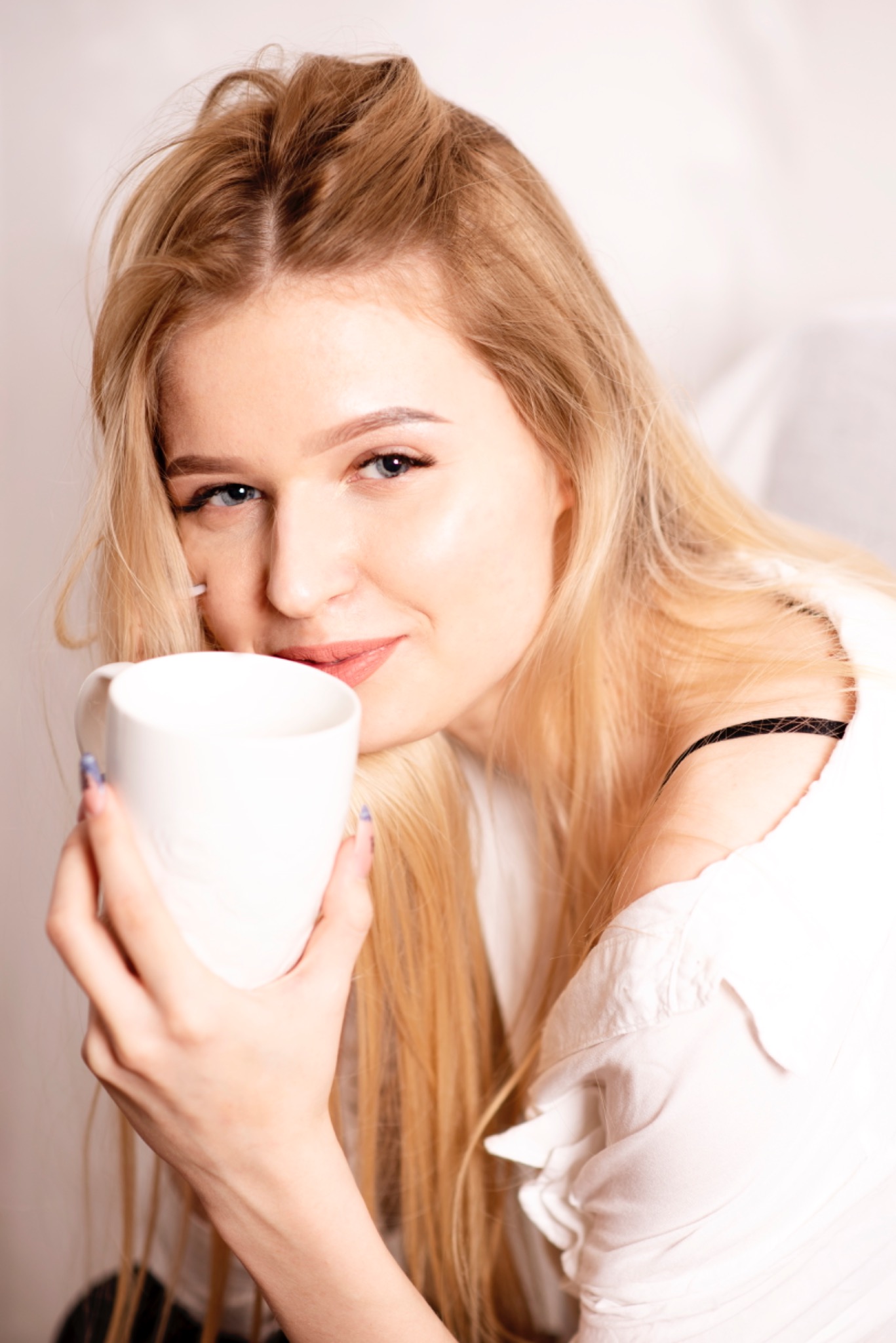 Uśmiechnięta blondynka w białej koszuli pije z białego kubka, patrząc w obiektyw.